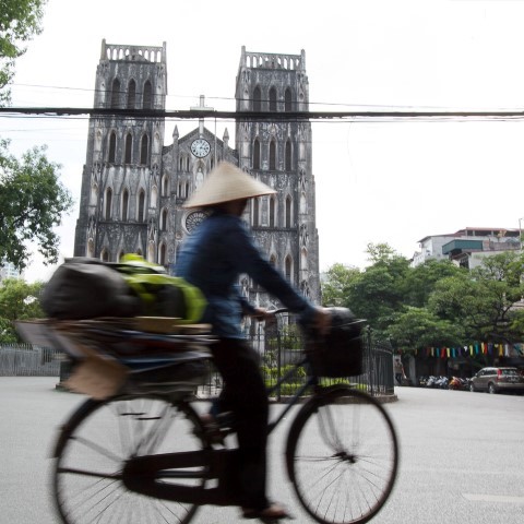 Photo of Vietnamese lady wearing conical hat cycling past the cathedral in Hanoi's French Quarter